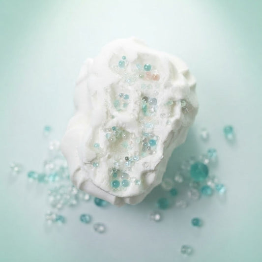 A white, textured bath bomb or soap piece with embedded clear and light blue beads, resting on a soft mint green background with scattered decorative beads around it.
