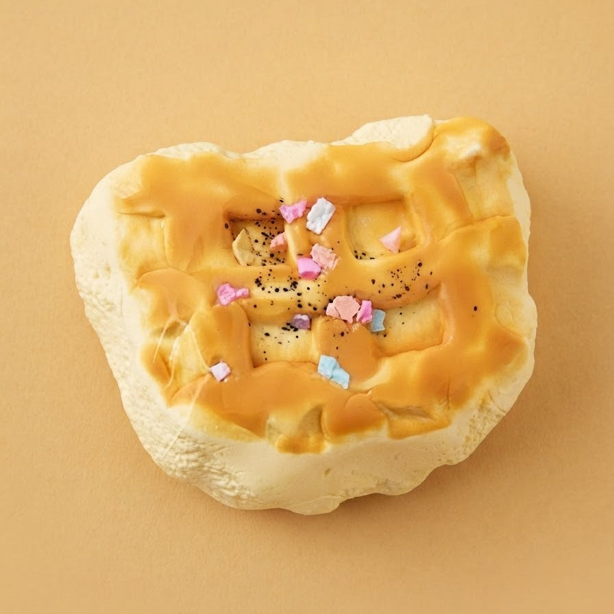 A novelty squishy toy shaped like a baked pastry or bread with a golden-brown crust, glossy orange glaze, and colorful sprinkles, shown on a solid tan background.