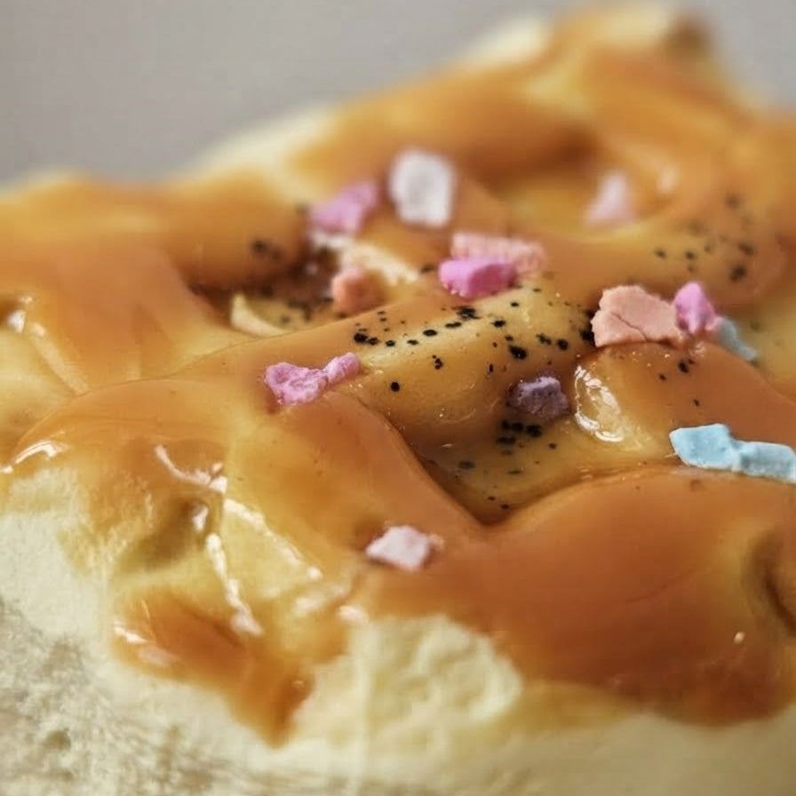 A close-up of a pastry or bread topping with glossy caramel-like drizzle, pink and blue candy-like sprinkles, and black speckles on a pale base.