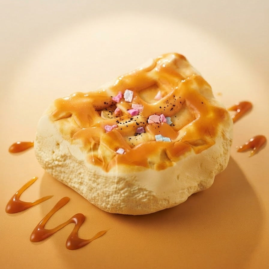 A dessert pastry topped with glossy caramel drizzle and small pink, blue, and white candy pieces, presented on a warm beige background with caramel sauce accents.