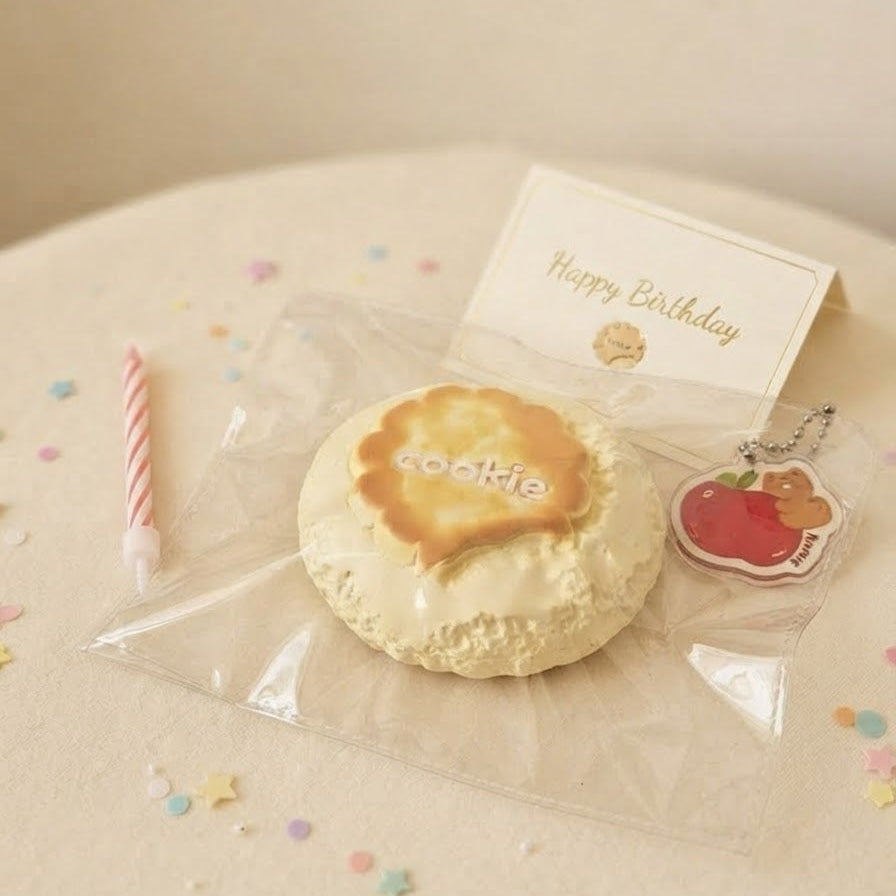 A packaged round cake-shaped item with a gold-foiled top and the word 'cookie' on it, placed on a beige surface with a small birthday candle, confetti, a card reading 'Happy Birthday,' and a small apple charm.