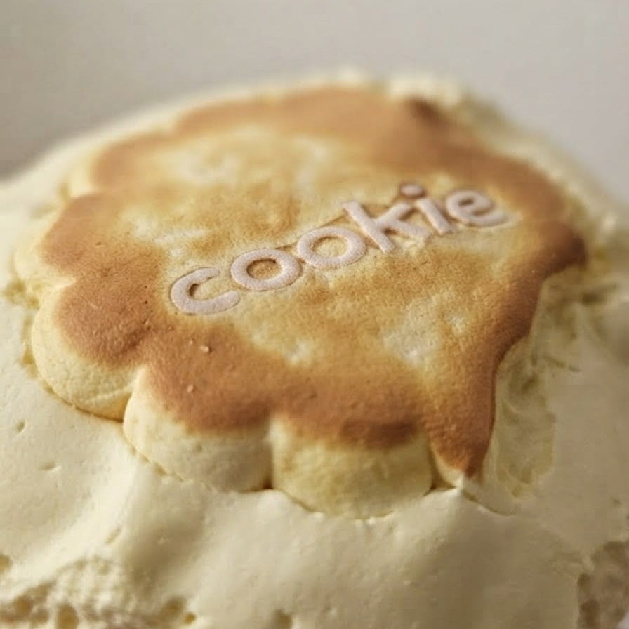 A close-up of a round, golden-brown baked cookie topped with thick white cream and the word 'cookie' spelled out in white icing, set against a soft, light beige background.