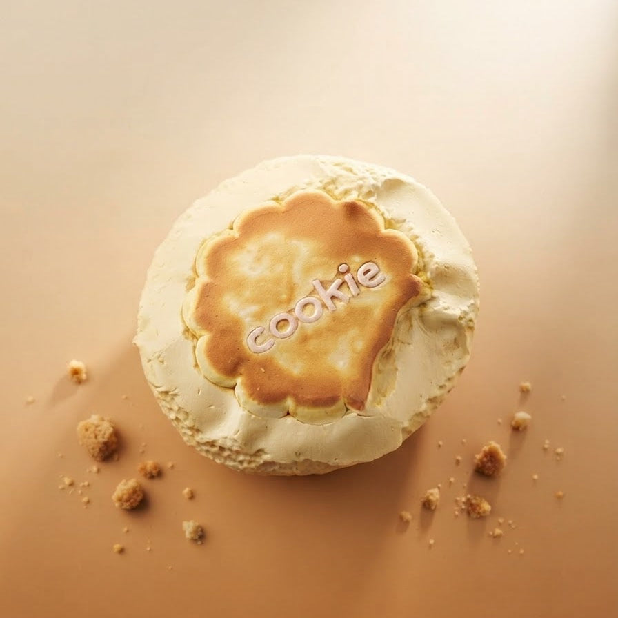 A round meringue-based dessert topped with a golden-brown caramelized center, photographed from above on a warm beige surface with a few crumbs nearby. The word 'cookie' is printed across the center.