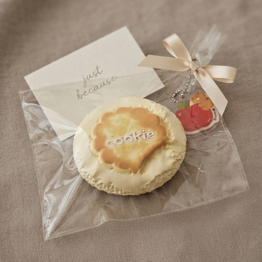 A round frosted cookie in clear plastic packaging with a cream ribbon and a small charm, placed on a beige fabric surface next to a card that reads 'just because.'.
