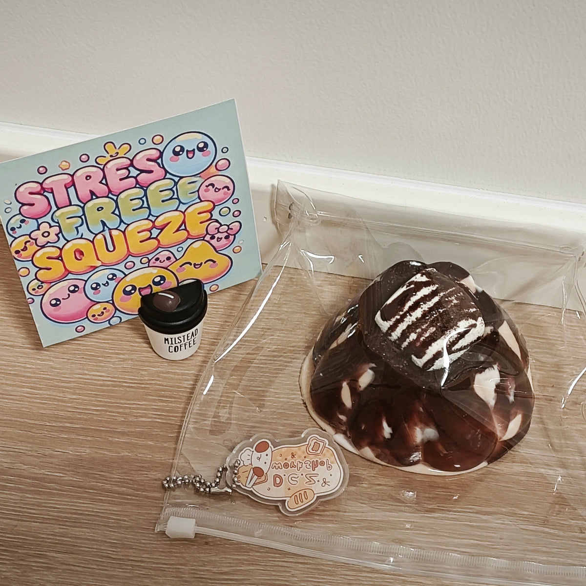 A chocolate-donut-shaped squishy toy sealed in a clear plastic bag, displayed on a light wood floor next to a pastel 'STRESS FREE SQUEEZE' card and a small cup accessory.