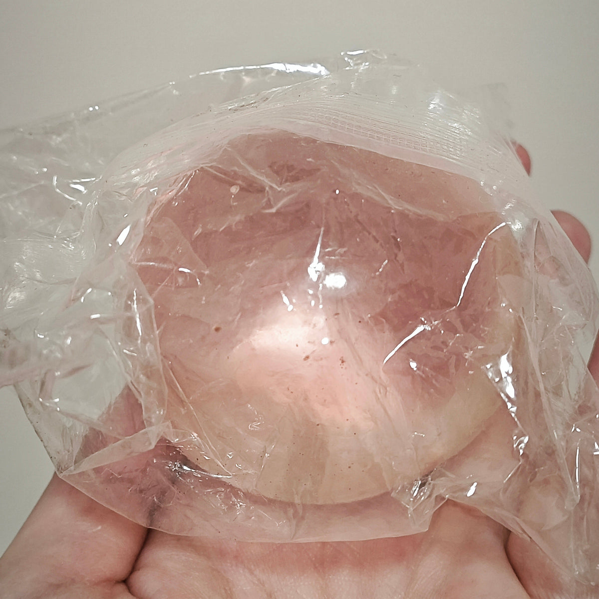 A hand holding a translucent, flesh-toned silicone breast prosthetic or training device wrapped in clear plastic against a plain light background.