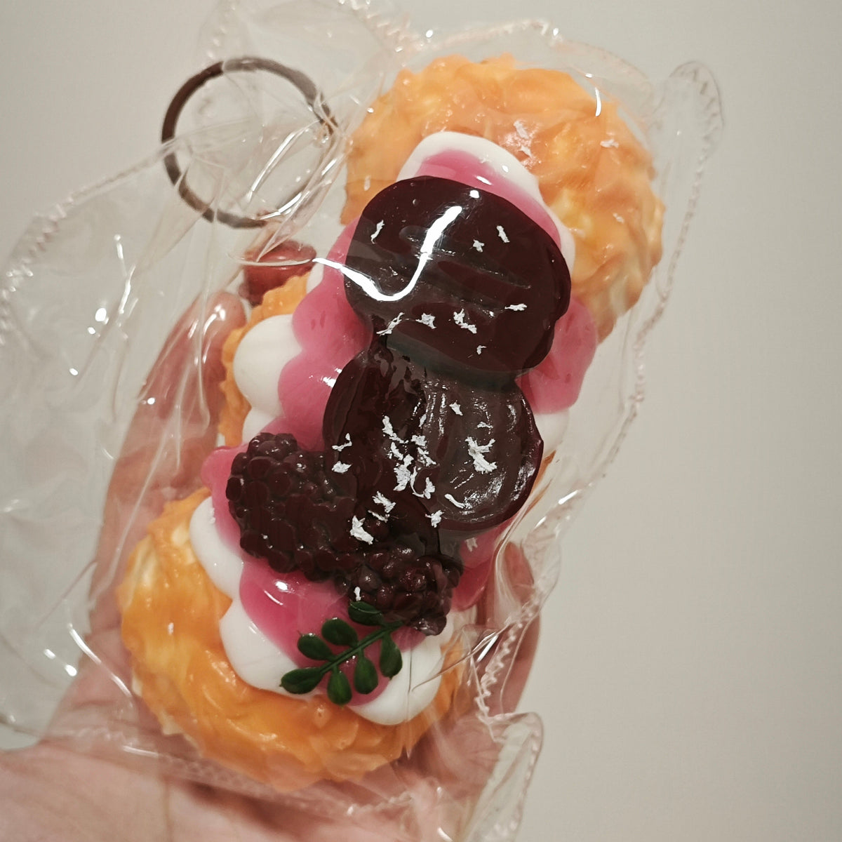 A hand holding a small, packaged novelty item shaped like a dessert, wrapped in clear plastic with a metal key ring attached. The dessert-style item has orange pastry-like sections, white and pink filling, dark brown chocolate-like drizzle, and a small green leaf detail, against a plain light background.