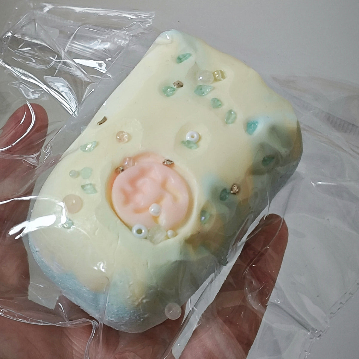 A handmade soap bar wrapped in clear plastic, held in a hand. The soap has a pale cream base with light blue and mint green marbling, a central pink circular detail, and small decorative beads.