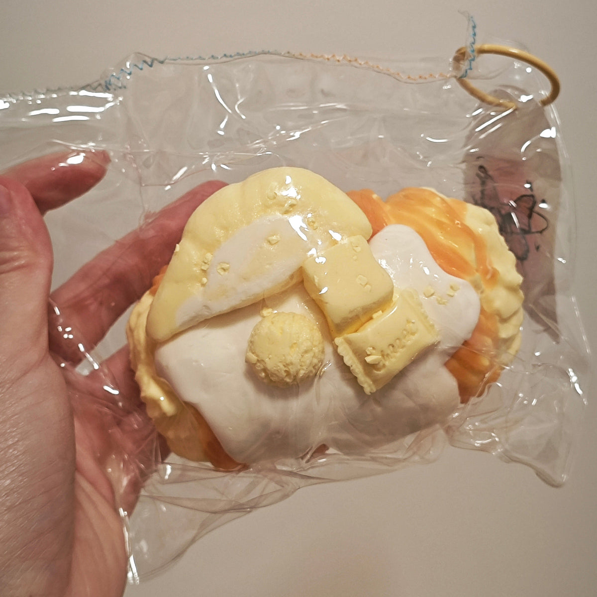 A hand holding a packaged novelty item shaped like a cream-colored angel with yellow hair and orange wings, sealed in clear plastic with a small gold ring at the top, against a plain light background.