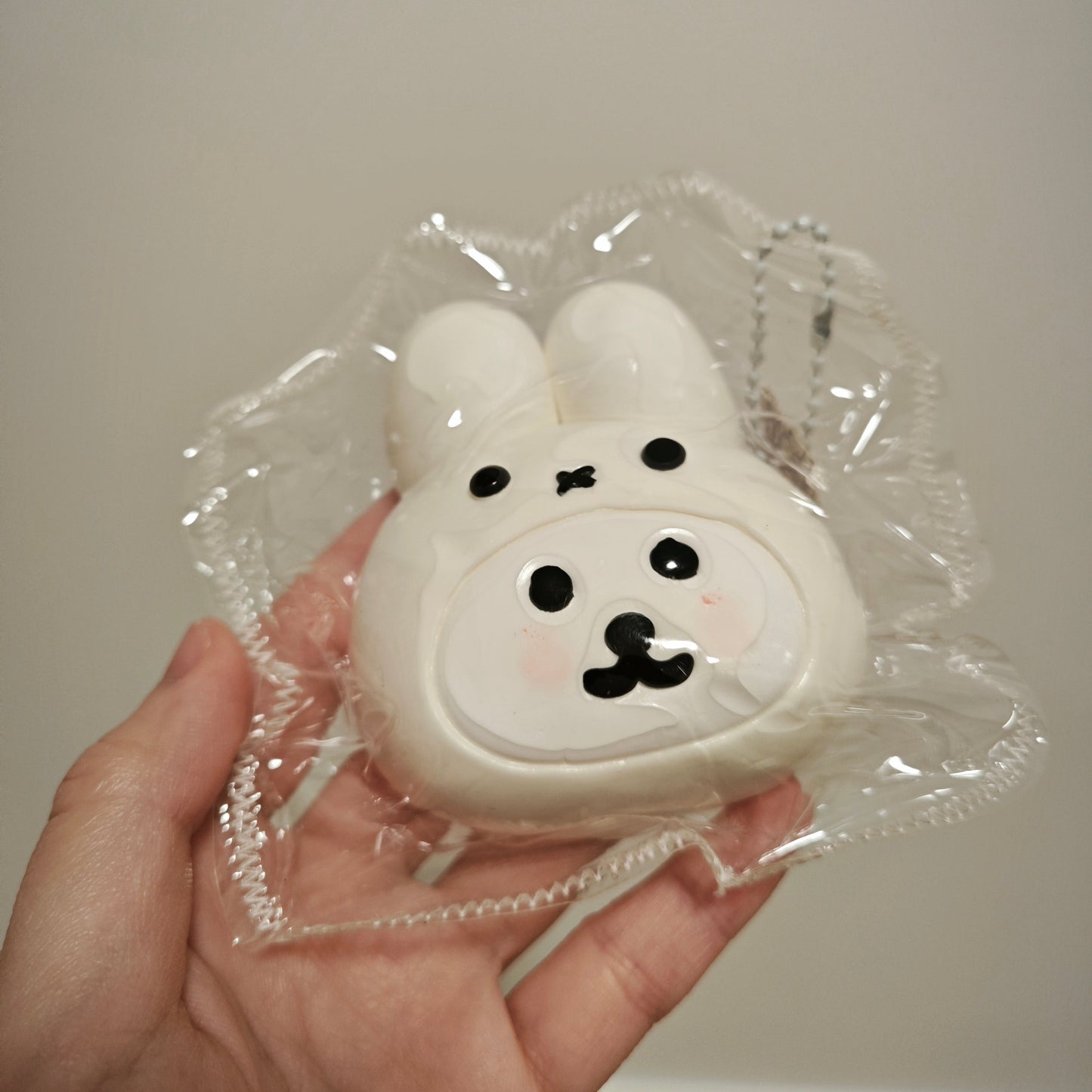 A hand holding a small white bunny-shaped accessory in clear plastic packaging against a plain light gray wall.