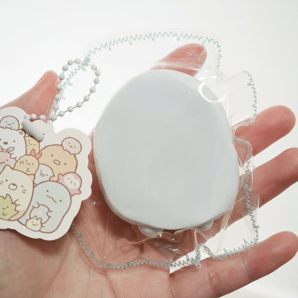 A hand holding a small white oval item in clear plastic packaging with a silver beaded chain and a cartoon character tag.