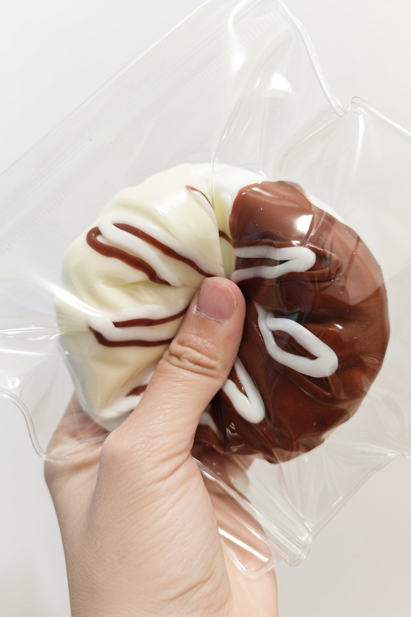 A hand holding a novelty donut-shaped item inside a clear plastic bag. The donut is split into two halves: one half is white with dark brown drizzle, and the other is dark brown with white drizzle, mimicking a chocolate-donut-flavored squishy or toy.