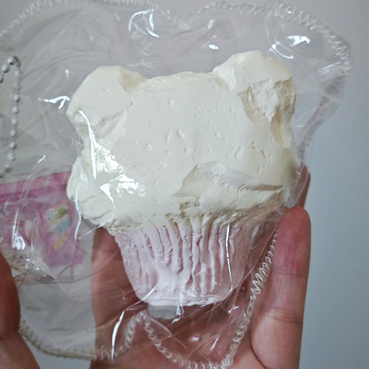 A hand holding a white frosted cupcake-shaped item inside a clear plastic bag with a scalloped edge.