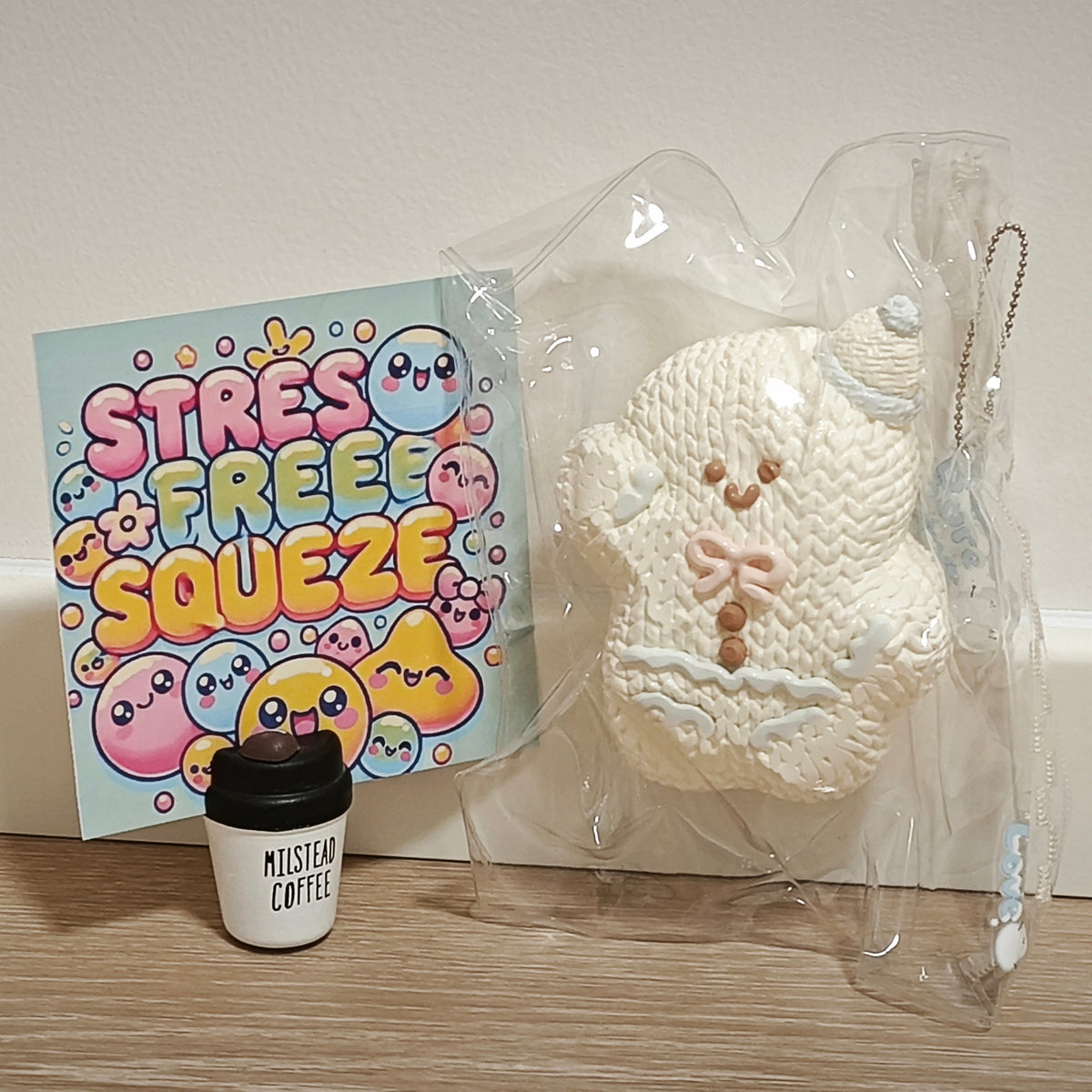 A cream-colored knitted bear-shaped squishy toy in clear plastic packaging, shown next to a colorful 'STRESS FREE SQUEEZE' card and a small black coffee cup on a wooden surface against a white wall.