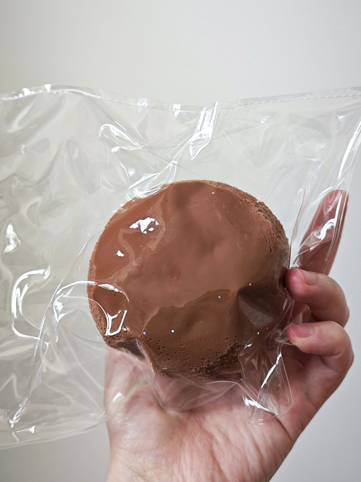 A hand holding a round chocolate brown item sealed in clear plastic packaging against a plain light background.