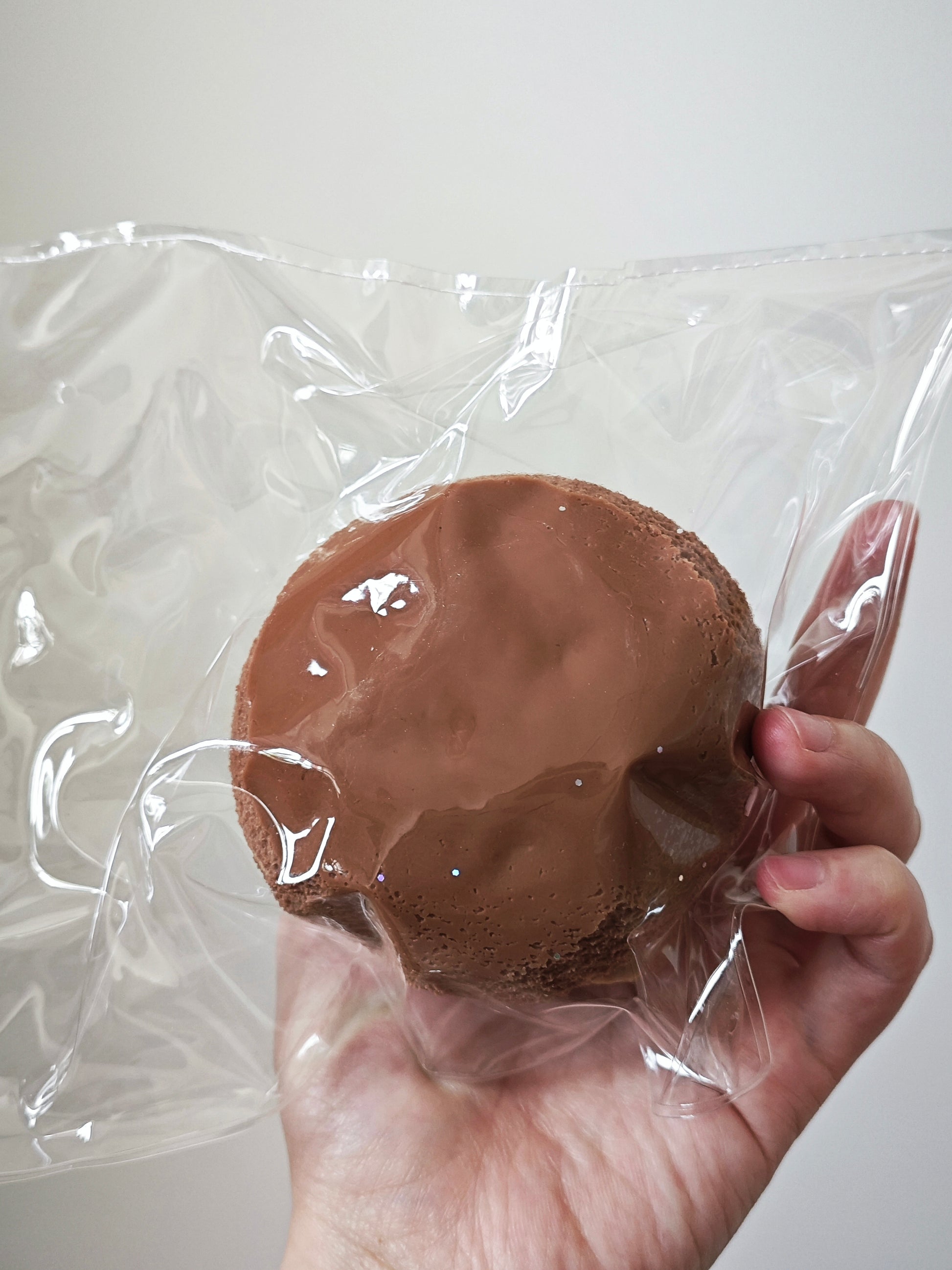 A hand holding a round chocolate brown item sealed in clear plastic packaging against a plain light background.