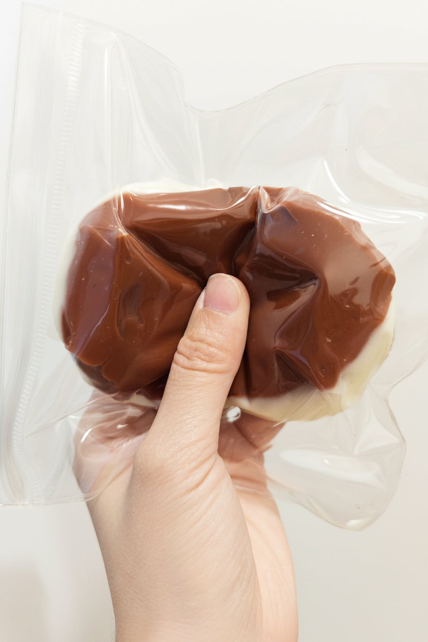 A hand holding a clear plastic bag containing a brown, crumpled, squishy object against a plain light background.