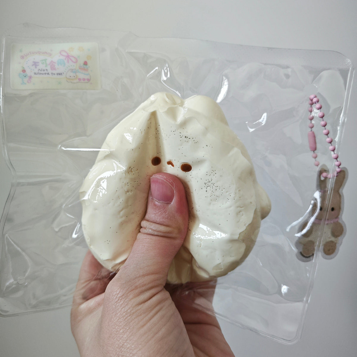 A hand holding a cream-colored, bunny-shaped squishy toy inside its clear plastic packaging, with a small pink-and-white keychain charm attached on the right side.