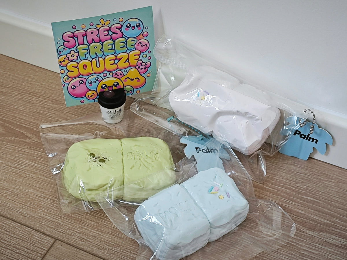A collection of pastel-colored squishies and accessories displayed on a light wood floor against a white wall. The set includes two rectangular squishies in light green and light blue inside clear plastic bags, a white squishy, a small blue keychain, a small black jar, and a colorful 'STRESS FREE SQUEEZE' card.
