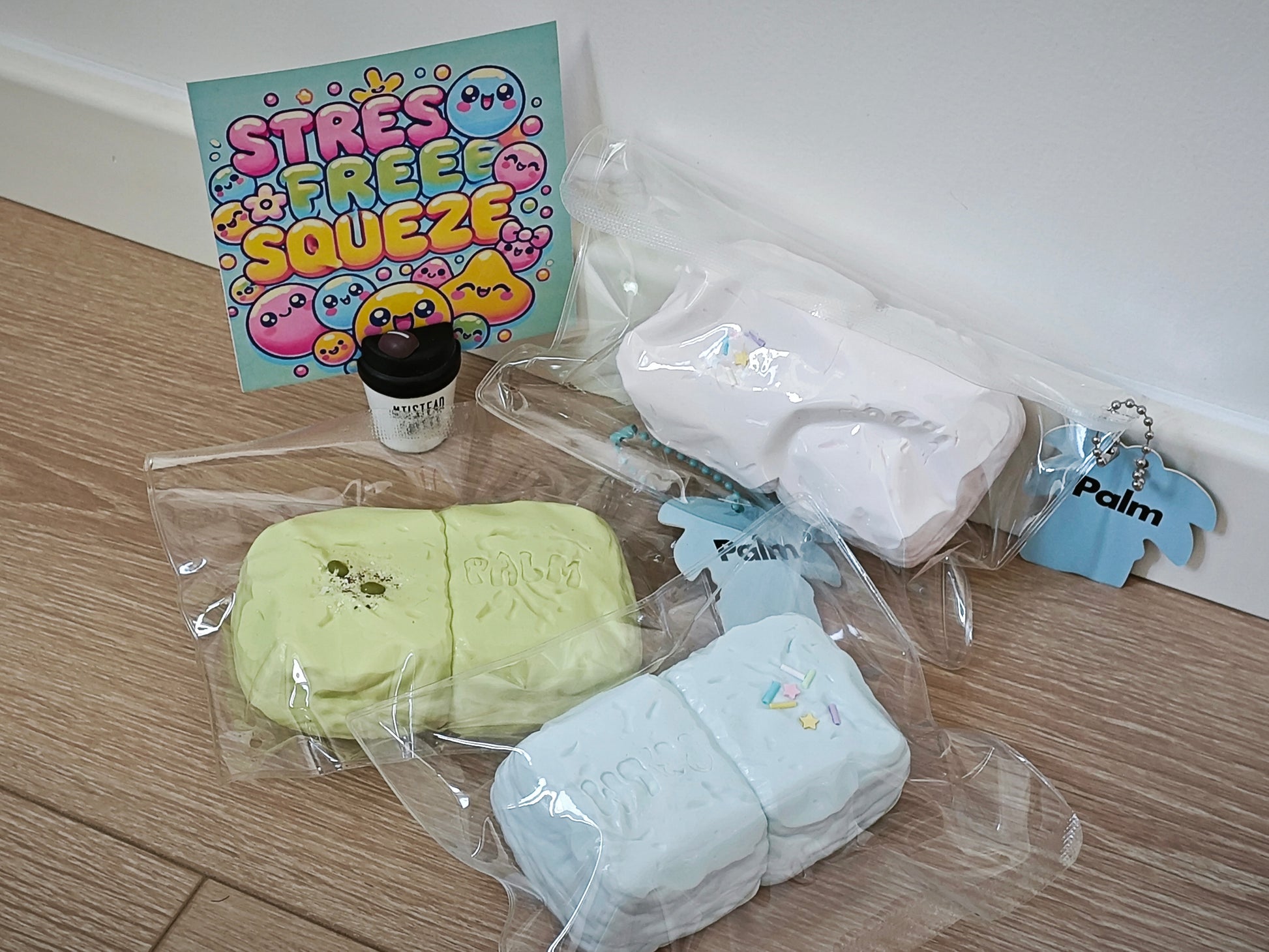 A collection of pastel-colored squishies and accessories displayed on a light wood floor against a white wall. The set includes two rectangular squishies in light green and light blue inside clear plastic bags, a white squishy, a small blue keychain, a small black jar, and a colorful 'STRESS FREE SQUEEZE' card.