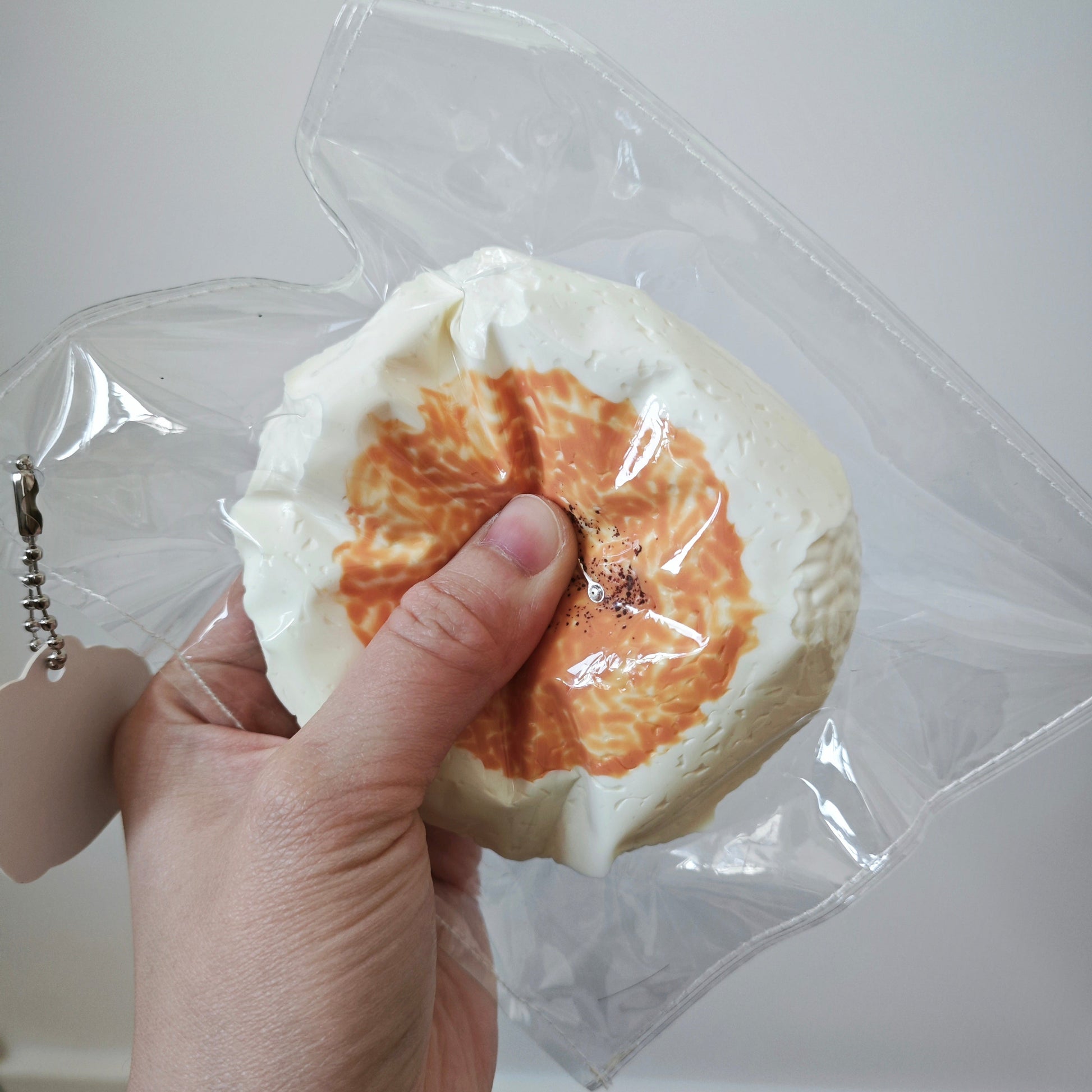 A hand holding a round, white, textured item with an orange citrus-patterned center, wrapped in clear plastic against a plain indoor wall.