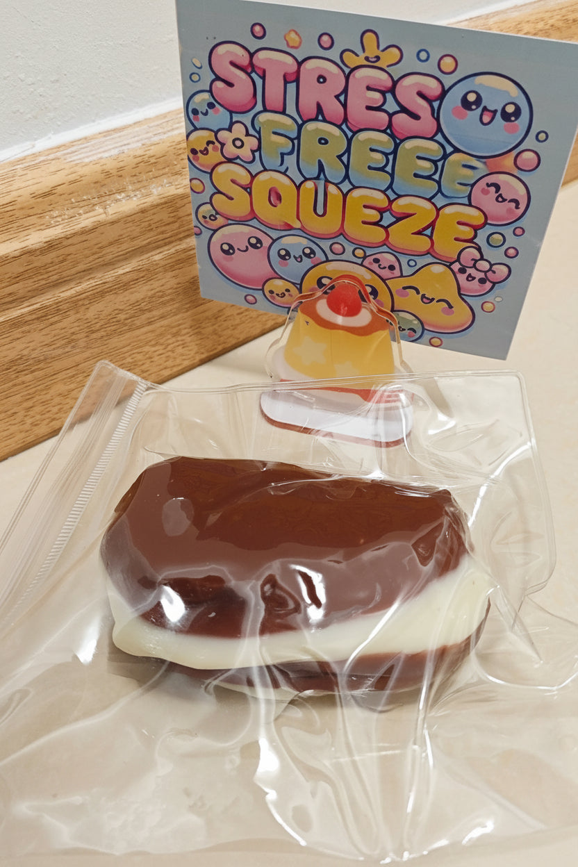 A packaged squishy toy shaped like a chocolate-covered sandwich with a white filling, sealed in a clear plastic bag, with a printed card behind it reading 'STRESS FREE SQUEEZE' and cartoon faces.