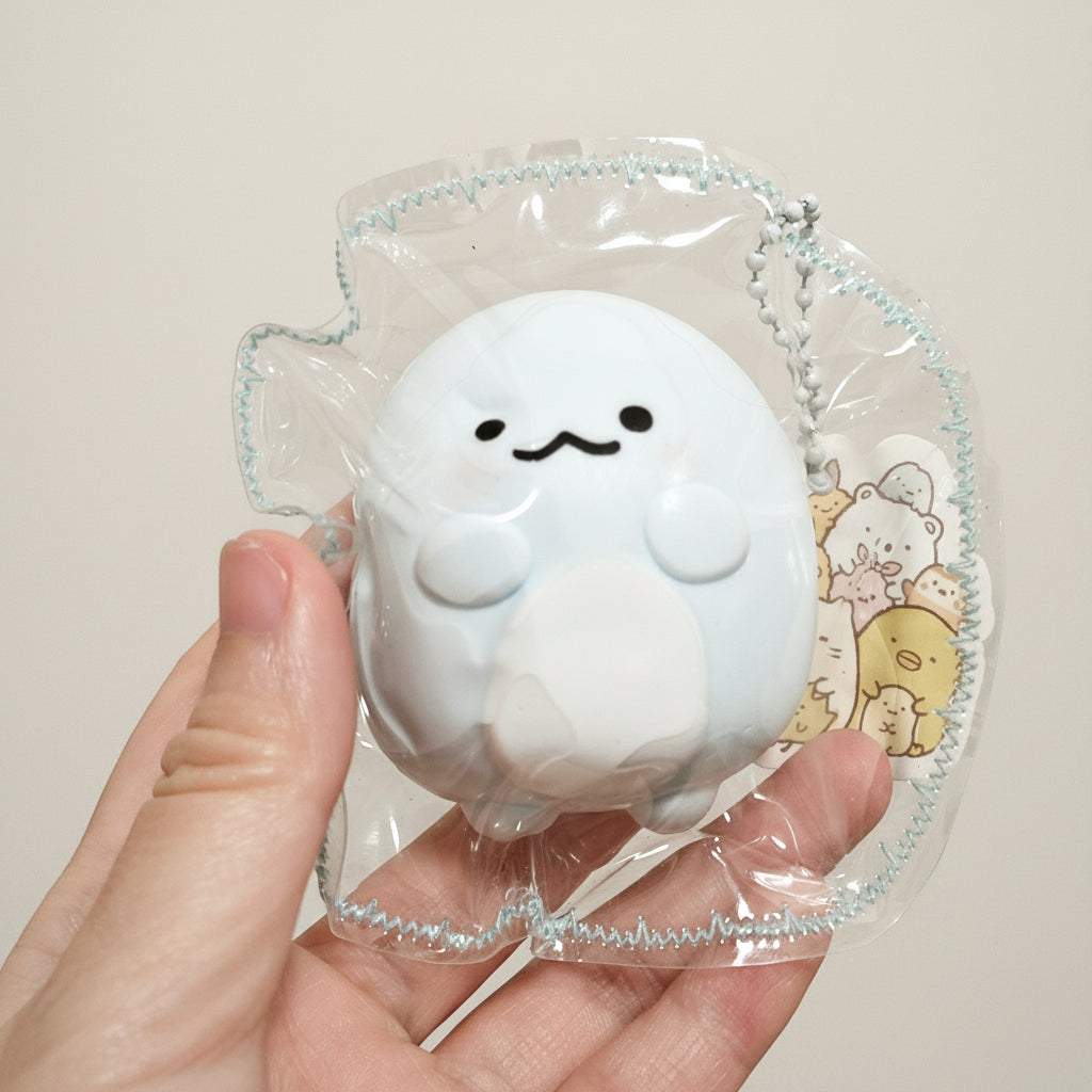 A hand holding a small white cartoon squishy toy inside a clear plastic bag with a silver chain, against a plain beige wall.