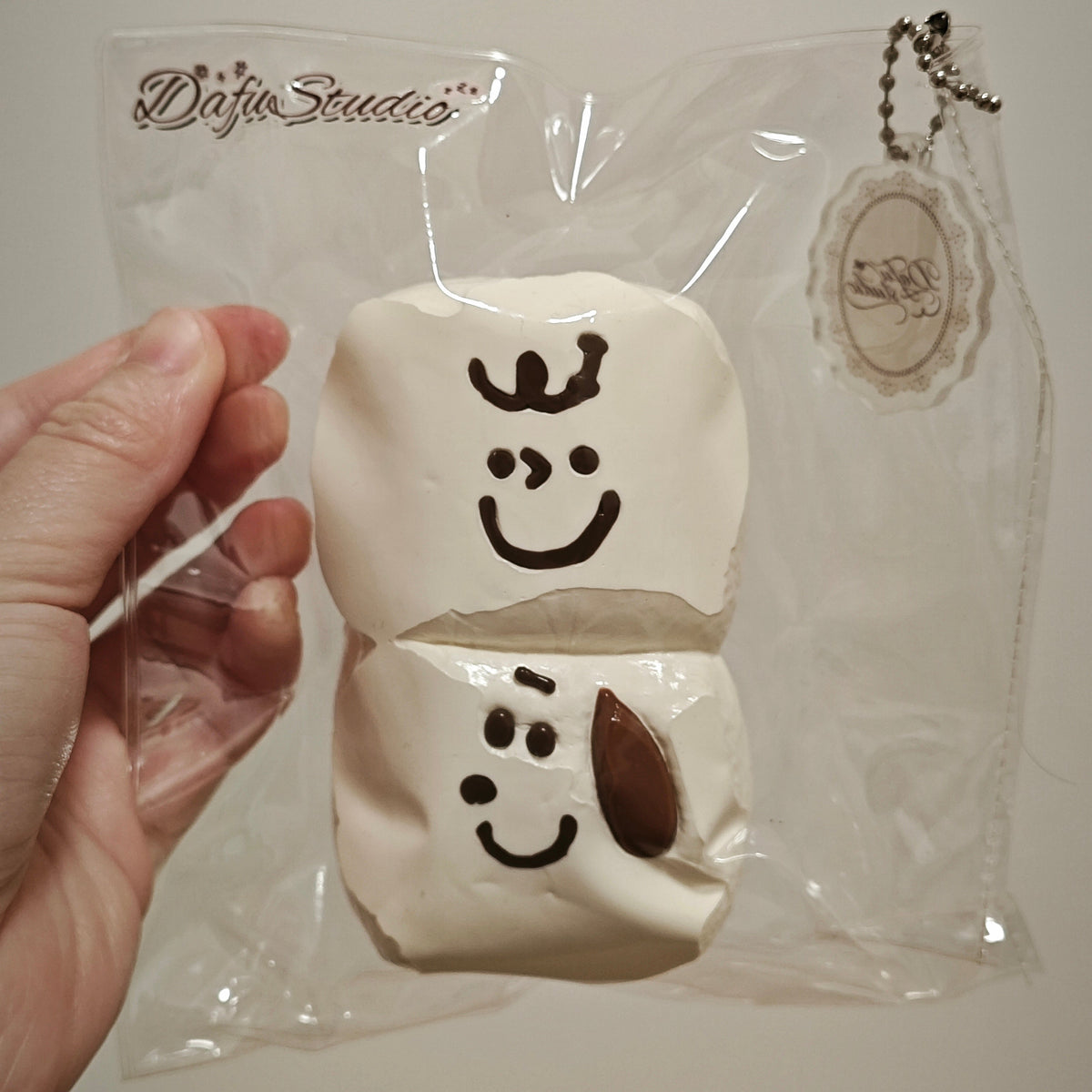A hand holds a packaged squishy toy featuring two stacked cream-colored marshmallow-like shapes with simple black face drawings. The item is sealed in a clear plastic bag with a visible 'Diafo Studios' logo and a hanging tag.