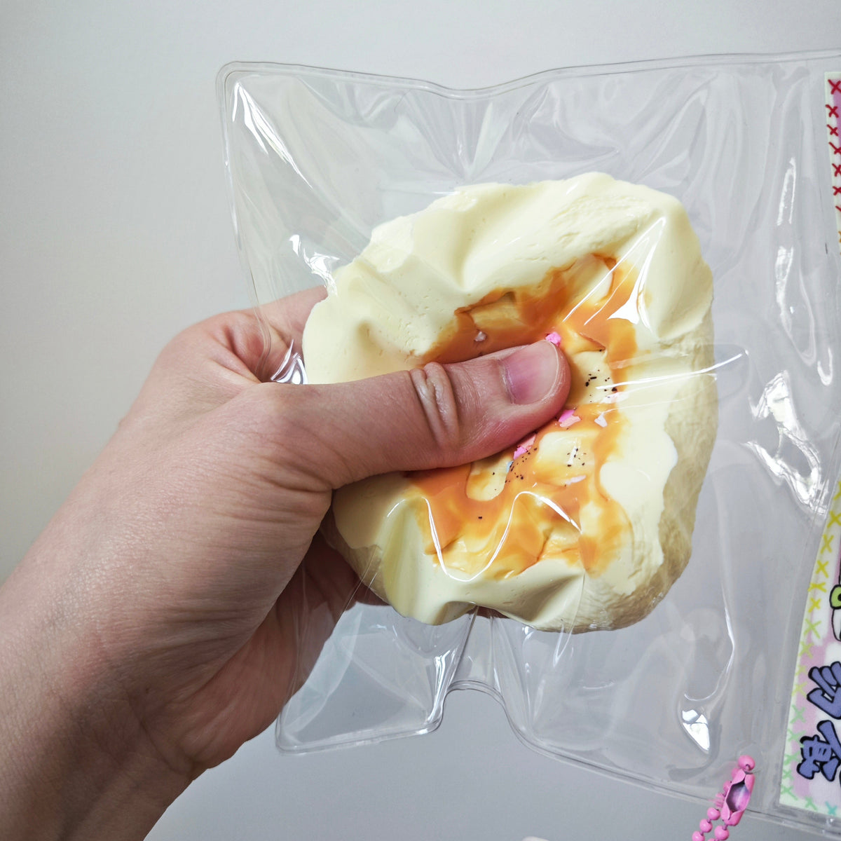 A hand holding a squishy toy shaped like a pastry or dumpling with orange sauce drizzle, packaged in a clear plastic bag against a plain light background.