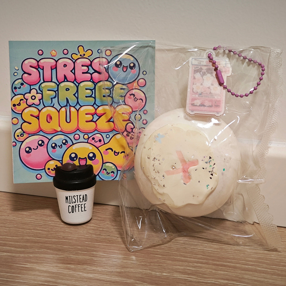 A cute squishy toy set on a wooden floor against a white baseboard. The set includes a large round cream-colored squishy with pink details and glitter, packaged in a clear plastic bag with a purple bead chain, a small white cup with a black lid labeled 'MISTLEAD COFFEE', and a colorful 'STRESS FREE SQUEEZE' card.