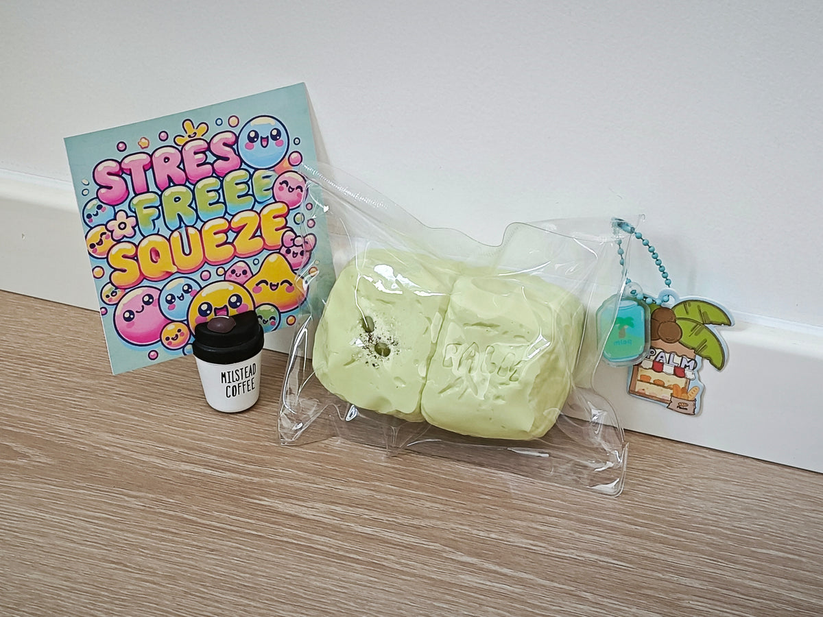 A pale yellow squishy toy in a clear plastic bag sits on a wood floor against a white wall, alongside a colorful 'STRESS FREE SQUEEZE' card, a small black jar labeled 'MISTY MIST COFFEE', and a small keychain.