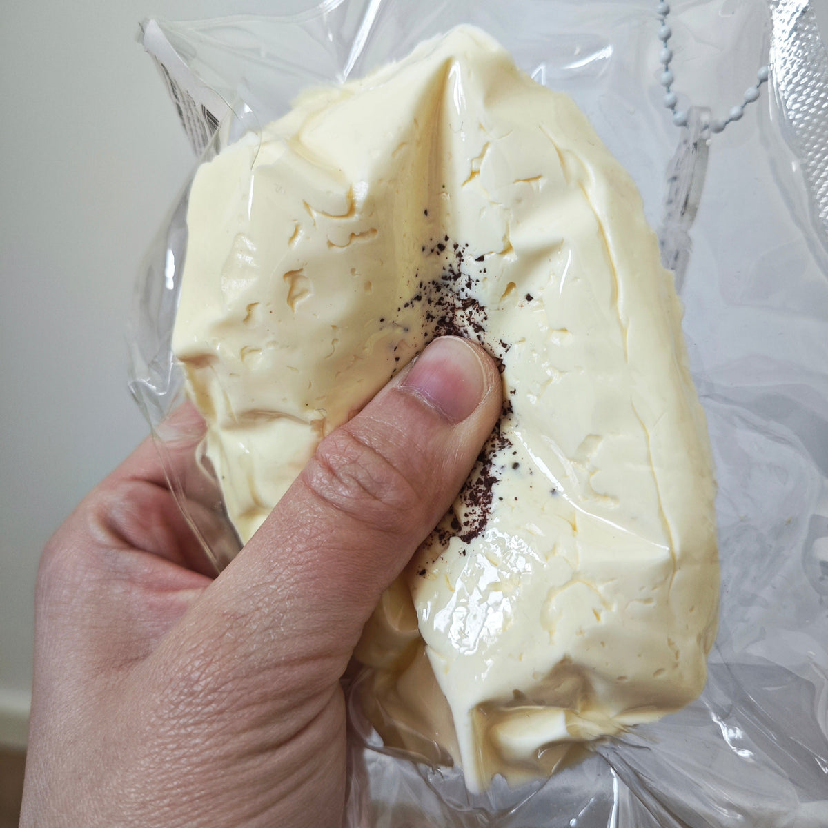 A hand holding a crumpled pale yellow block or puck-shaped item wrapped in clear plastic, with a small amount of dark powder sprinkled on top.