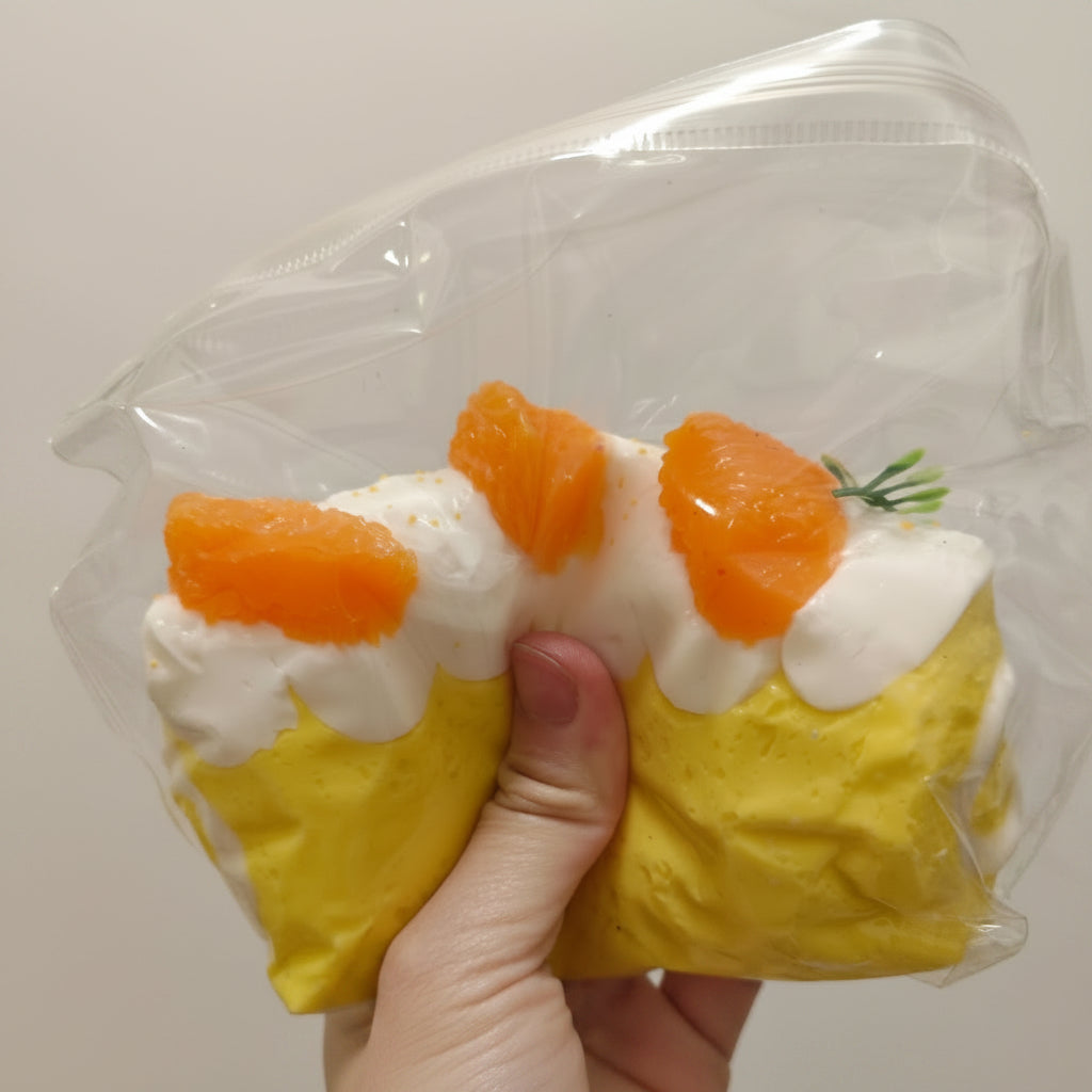 A hand holding a clear plastic bag containing a yellow and white novelty item decorated with orange slices and a small green sprig, against a plain light background.