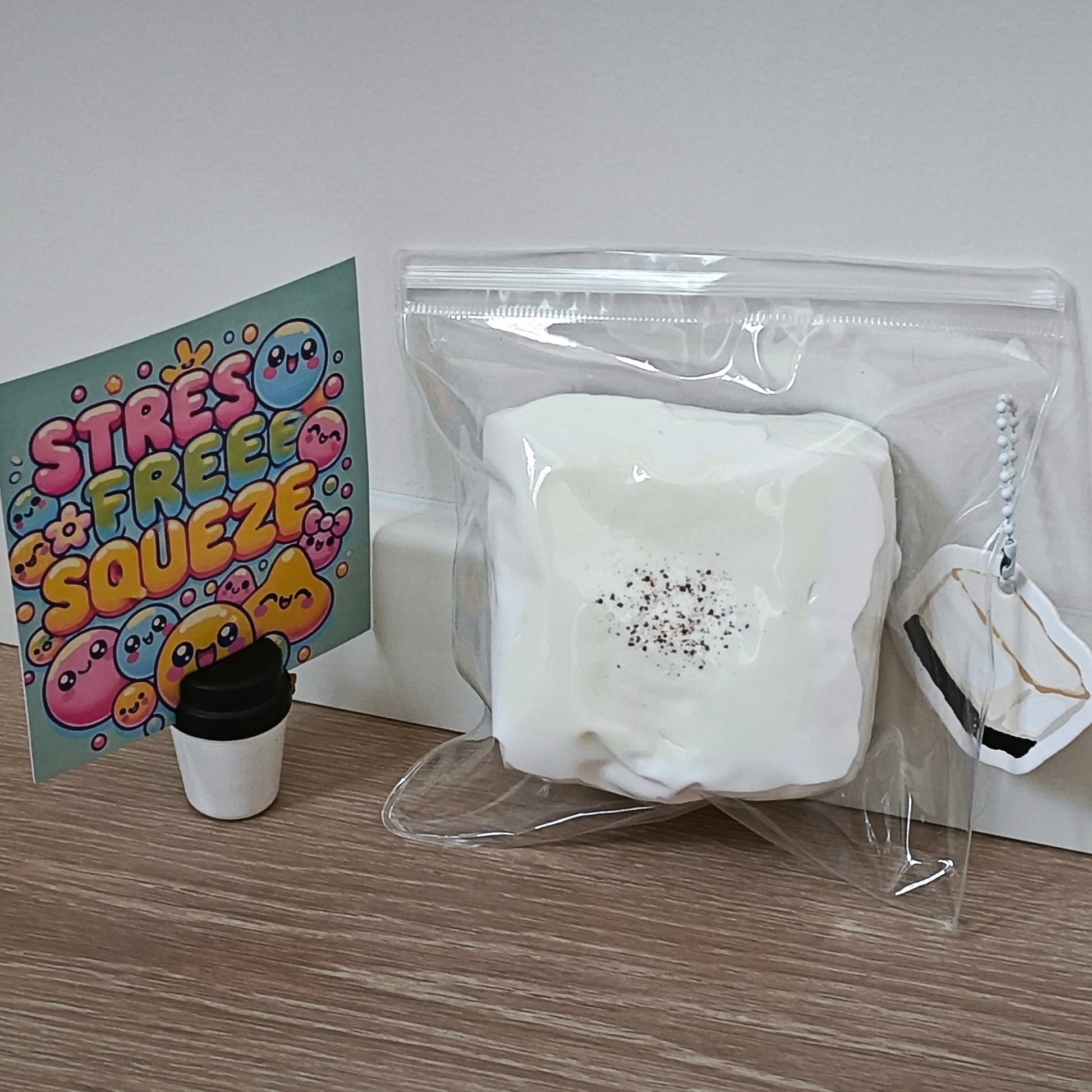 A white squishy toy packaged in a clear zip bag, displayed on a wooden surface next to a colorful 'STRESS FREE SQUEEZEE' card and a small black-and-white cup-shaped accessory.