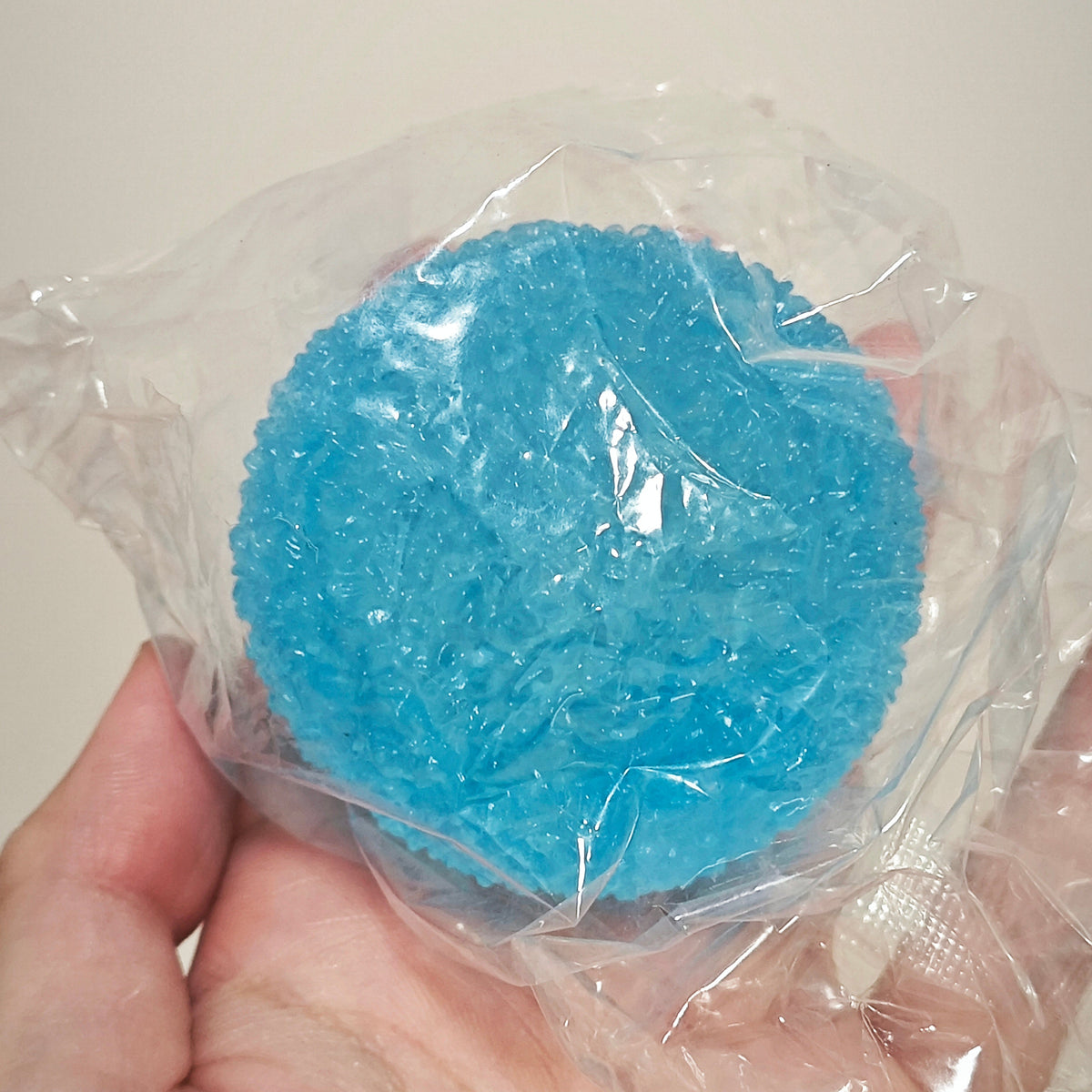 A hand holding a round, bright blue textured item wrapped in clear plastic against a plain light beige background.