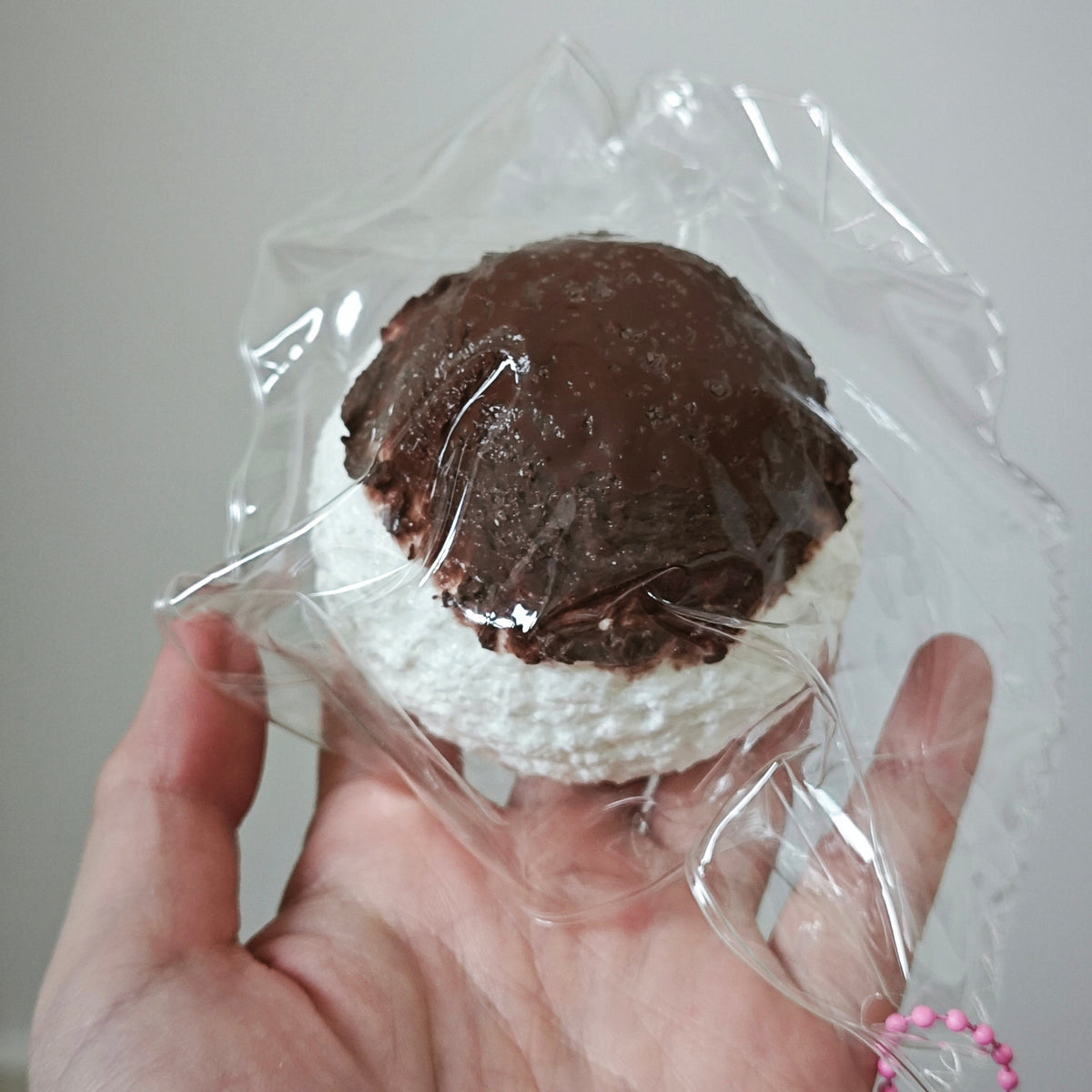 A hand holding a small round novelty item wrapped in clear plastic. The item has a dark brown top layer resembling chocolate frosting or a crust and a white textured bottom layer resembling marshmallow or cotton.