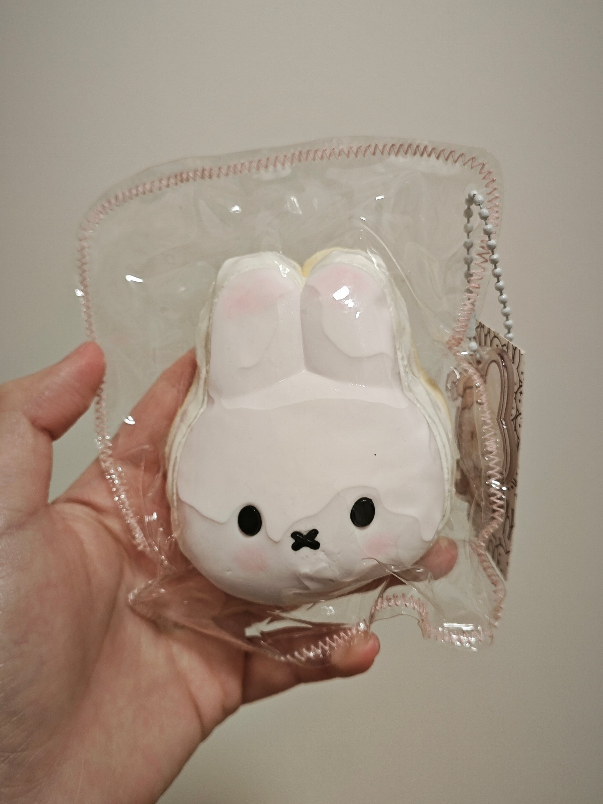 A hand holding a white bunny-shaped squishy toy inside a clear plastic bag with a pink stitched border and a silver chain.