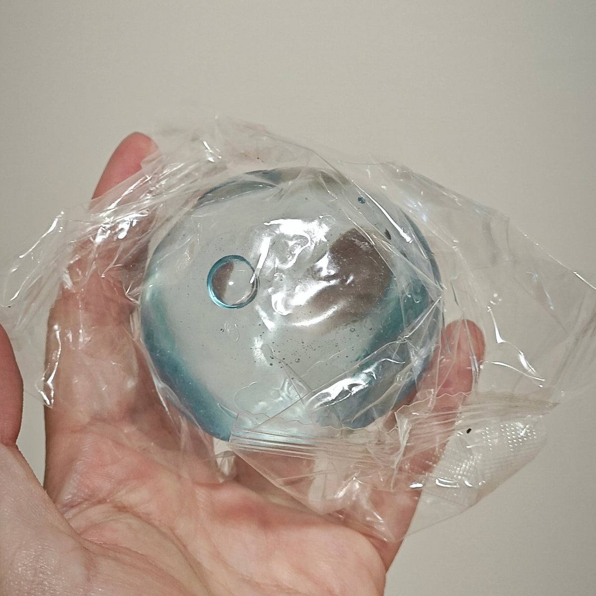 A hand holding a small round, clear plastic-wrapped item with a blue tint and a circular cutout, against a plain light background.