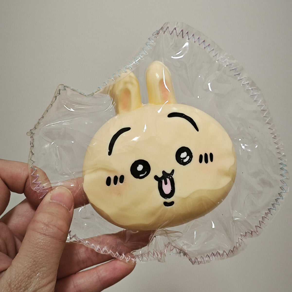 A hand holding a small, round, pale yellow cartoon bunny face toy inside a crinkled clear plastic bag against a plain light gray background.
