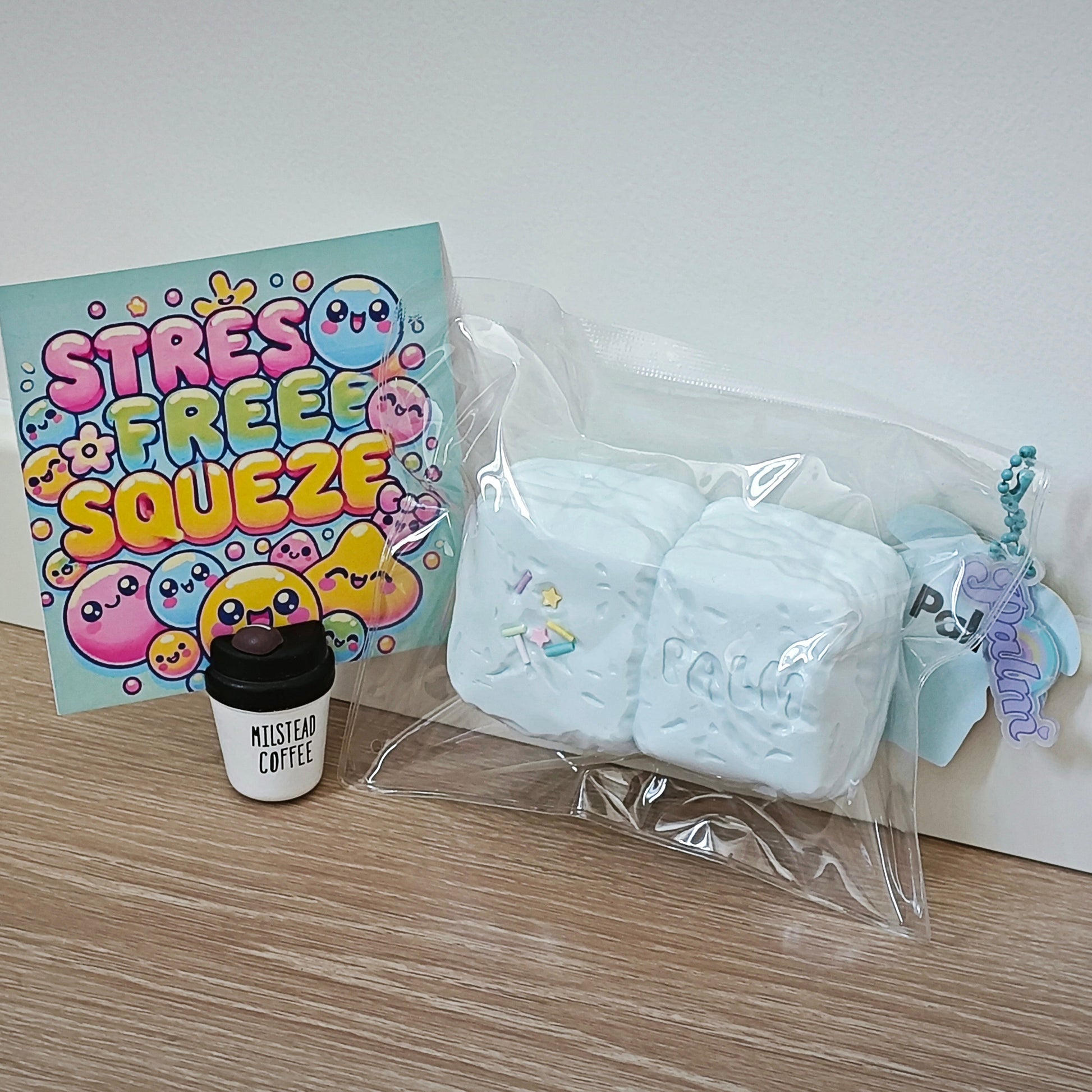 A set of stress-free self-care items arranged on a light wood floor against a white wall: a pastel 'STRESS FREE SQUEEZE' card, a small black-and-white 'MISTYAN COFFEE' cup, and a clear bag containing two pale blue squishy marshmallow-like pieces with a blue keychain charm.
