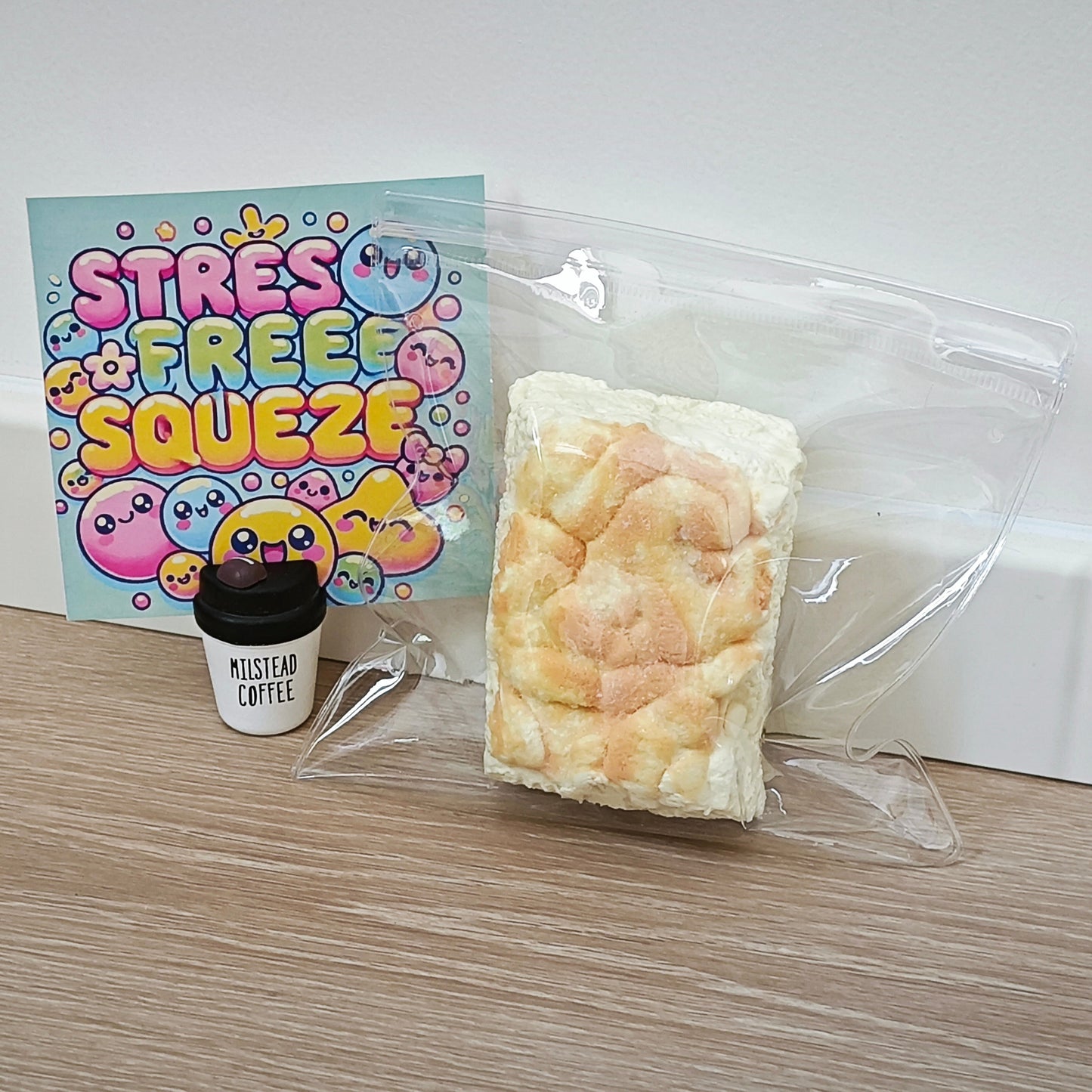 A packaged squishy bread toy sits on a wood surface next to a small coffee cup and a colorful 'STRESS FREE SQUEEZE' card. The product is sealed in a clear plastic bag and appears to be a rectangular bread-like toy with a pale cream surface and peachy baked areas.