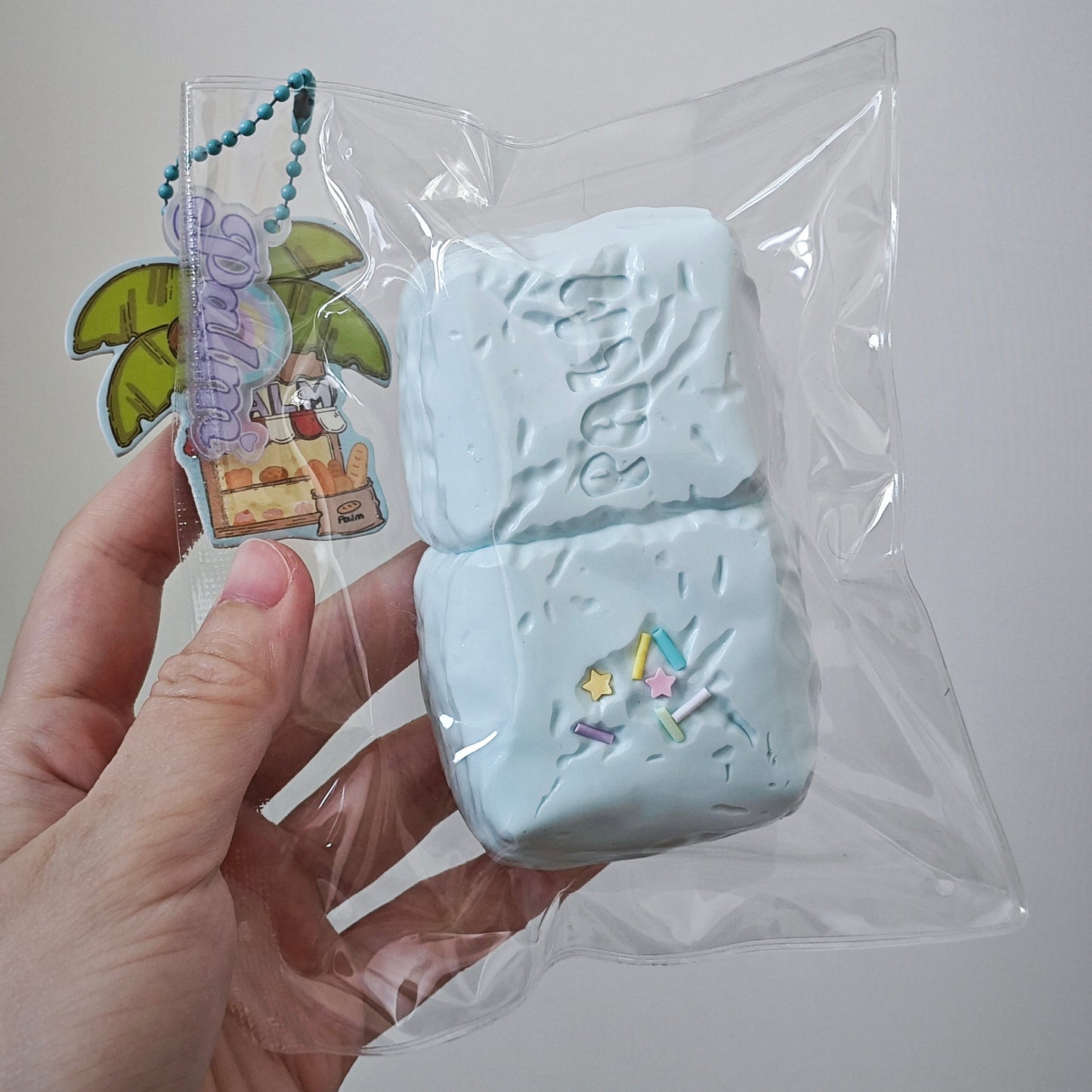 A hand holding a clear plastic bag containing a light blue textured rectangular item, with a small colorful keychain charm attached. The background is a plain light gray wall.