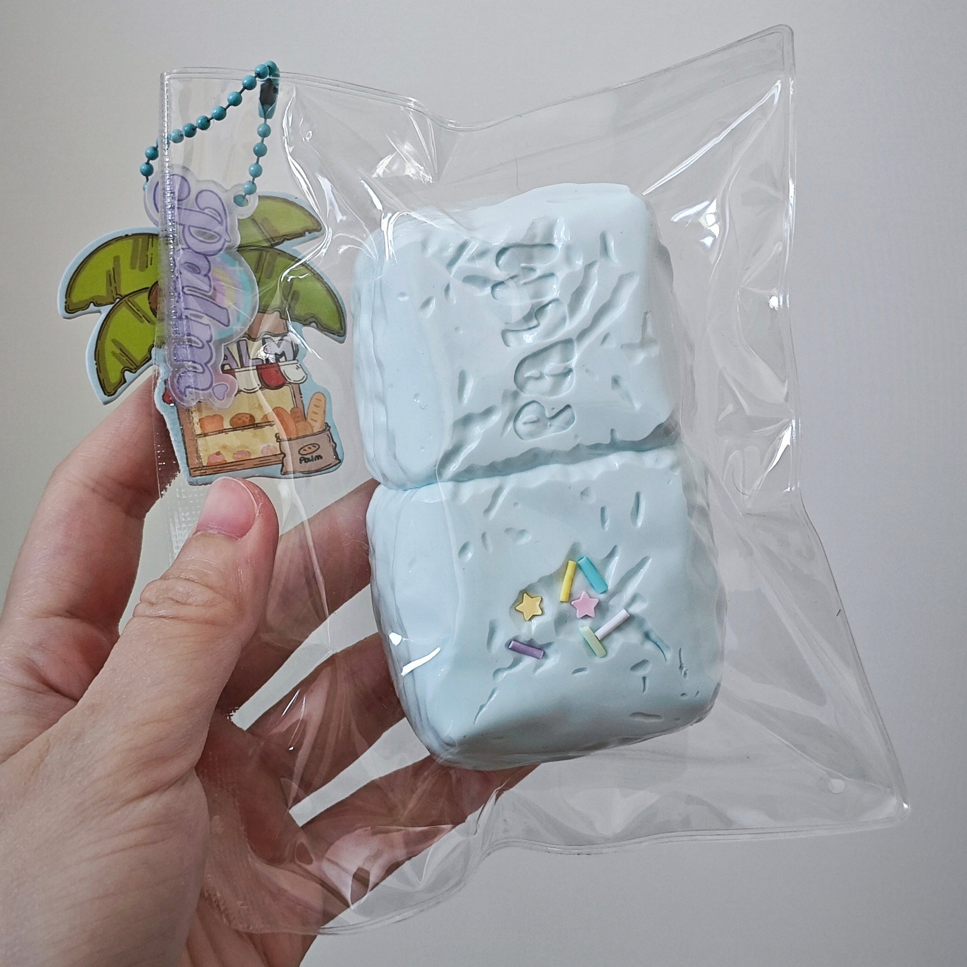 A hand holding a clear plastic bag containing a light blue textured rectangular item, with a small colorful keychain charm attached. The background is a plain light gray wall.