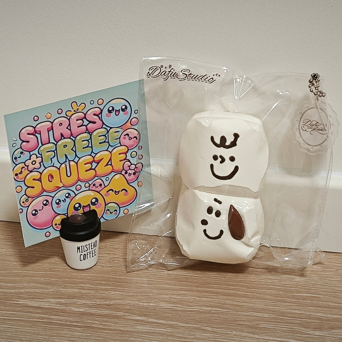 A white two-tier squishy toy with cartoon faces, shown in its clear plastic packaging next to a small coffee cup accessory and a colorful 'Stress Free Squeeze' card on a wooden surface.
