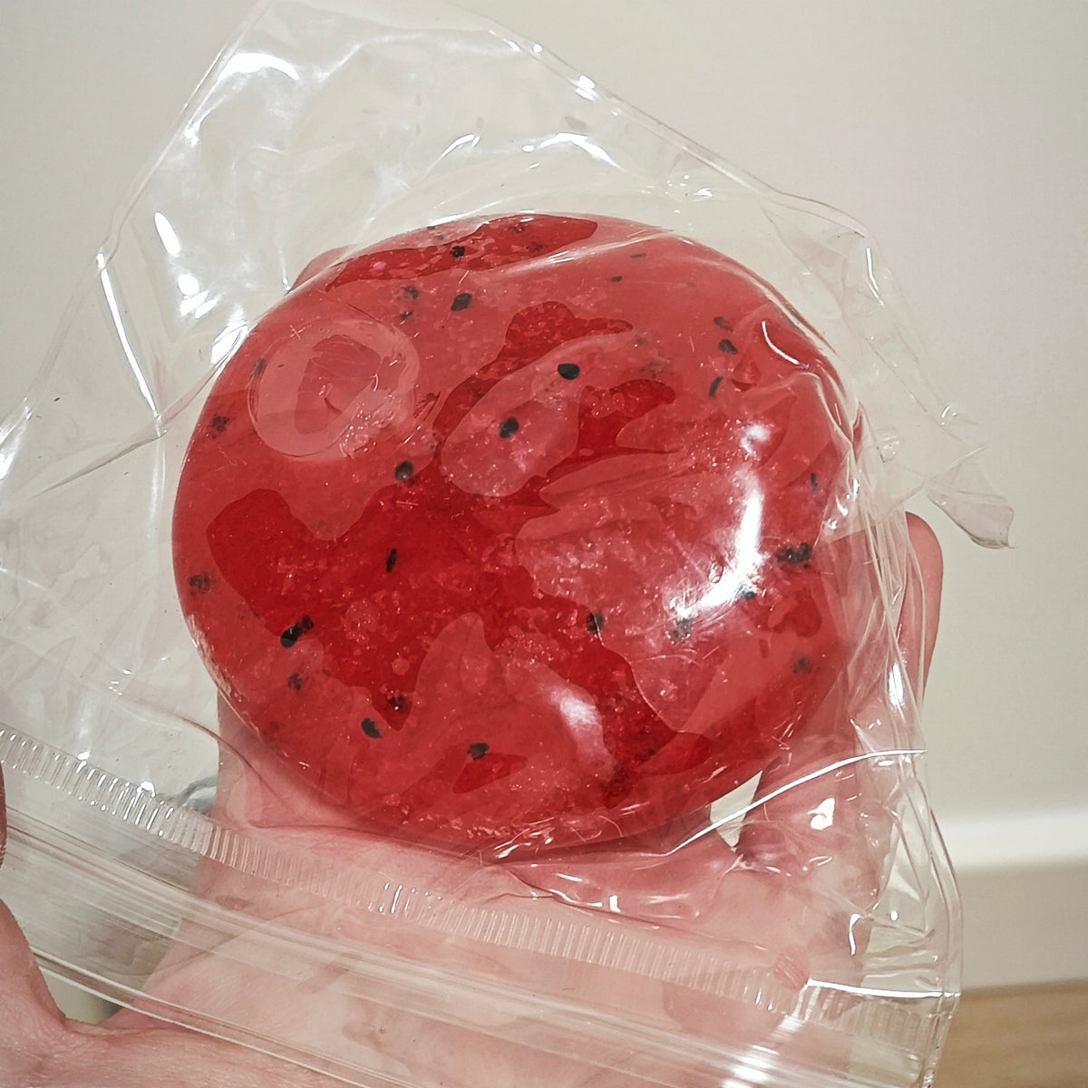 A red, round, gel-like or dough-like product with small black specks, sealed in a clear plastic bag and held in a person's hand against a plain indoor background.