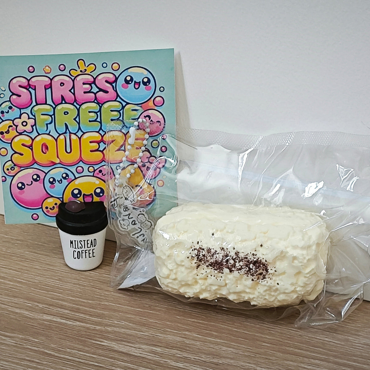 A packaged white textured squishy toy with dark brown sprinkles, shown next to a small black-and-white cup labeled 'MISTLEND COFFEE' and a colorful card that reads 'STRESS FREE SQUEEZE!' on a light wood floor against a white wall.