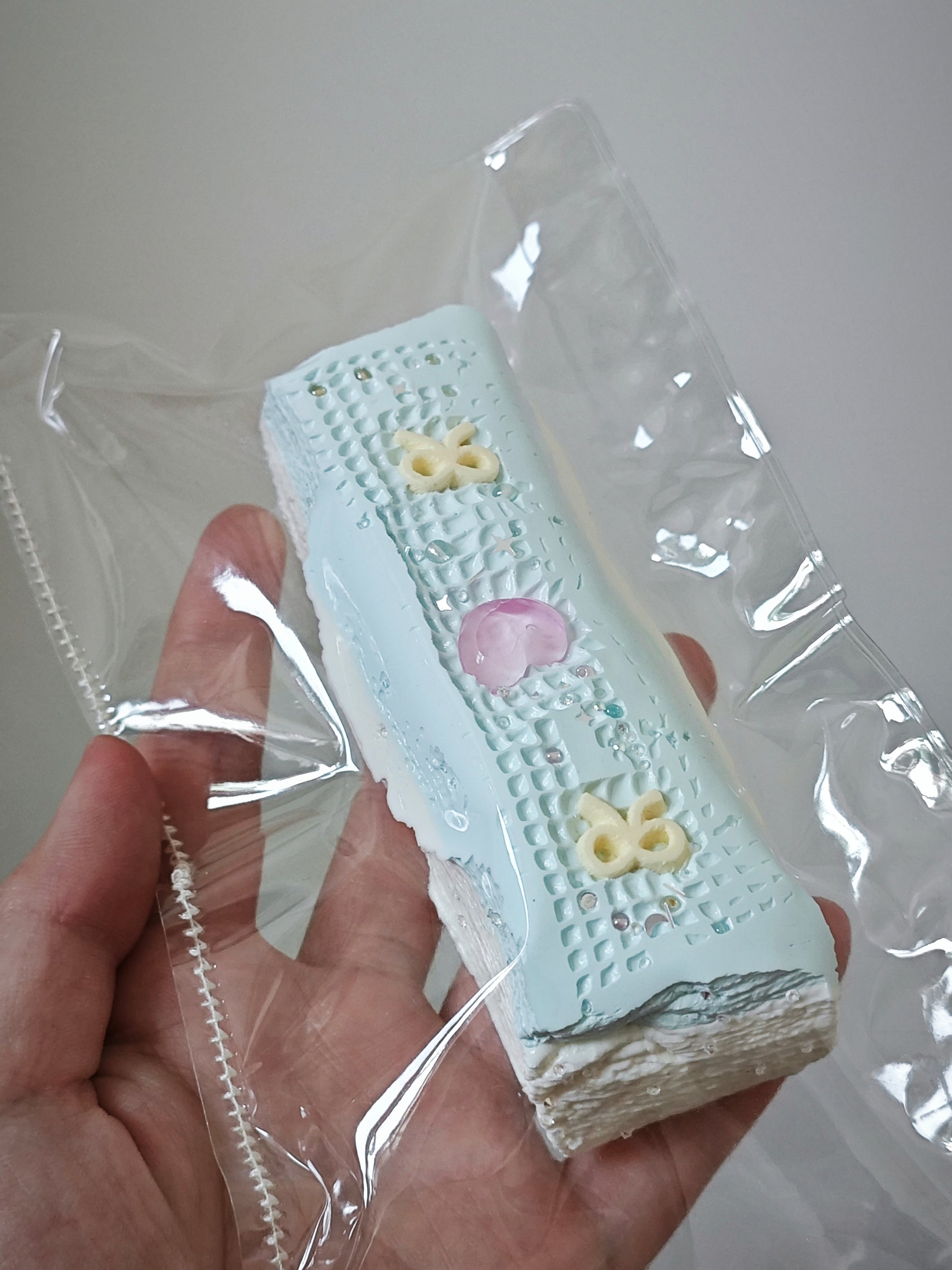 A hand holding a small rectangular pastel item wrapped in clear plastic. The item appears light blue with a textured pattern, a pink heart detail, and small yellow decorative accents.