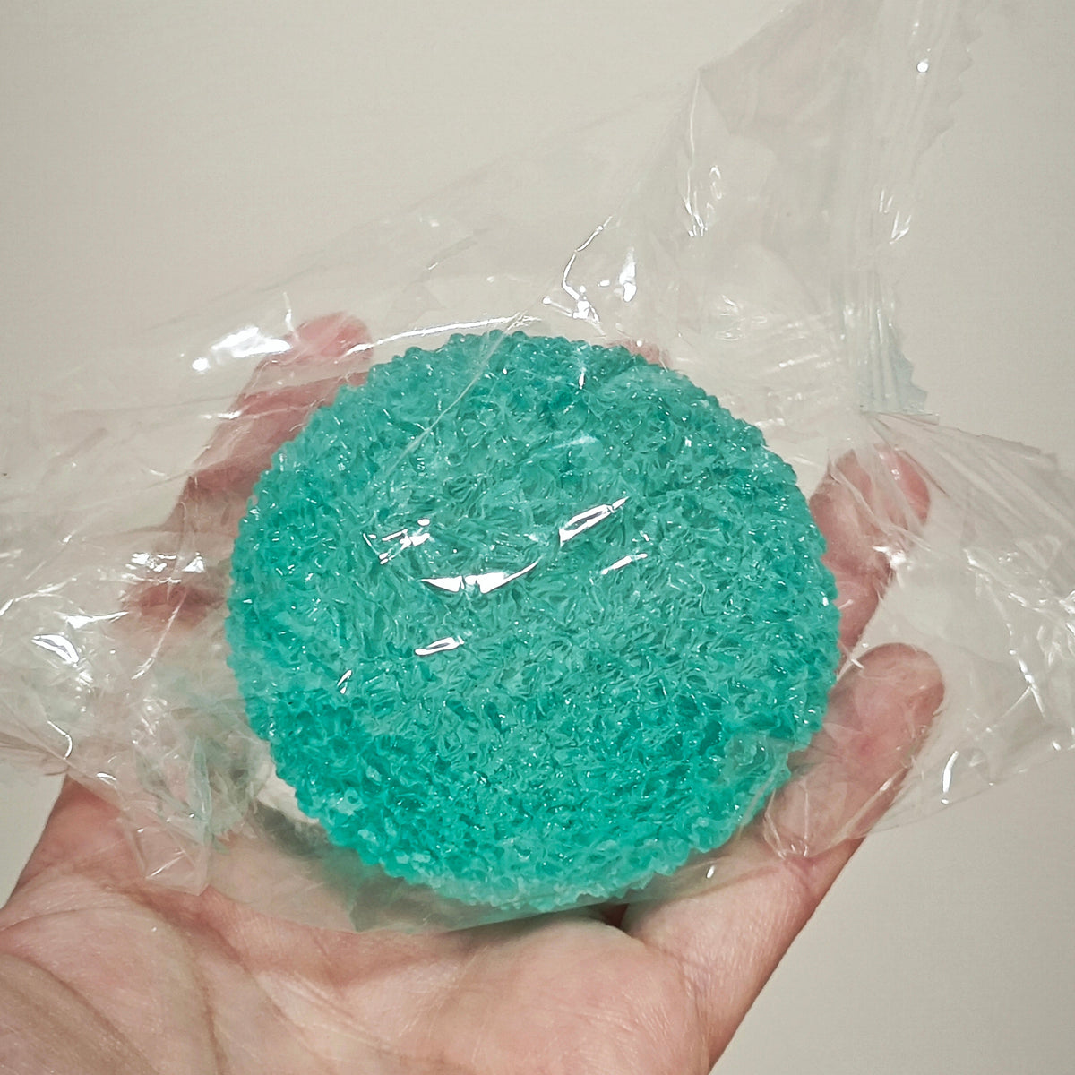 A hand holding a round, turquoise green scrubber or sponge wrapped in clear plastic against a plain light background.