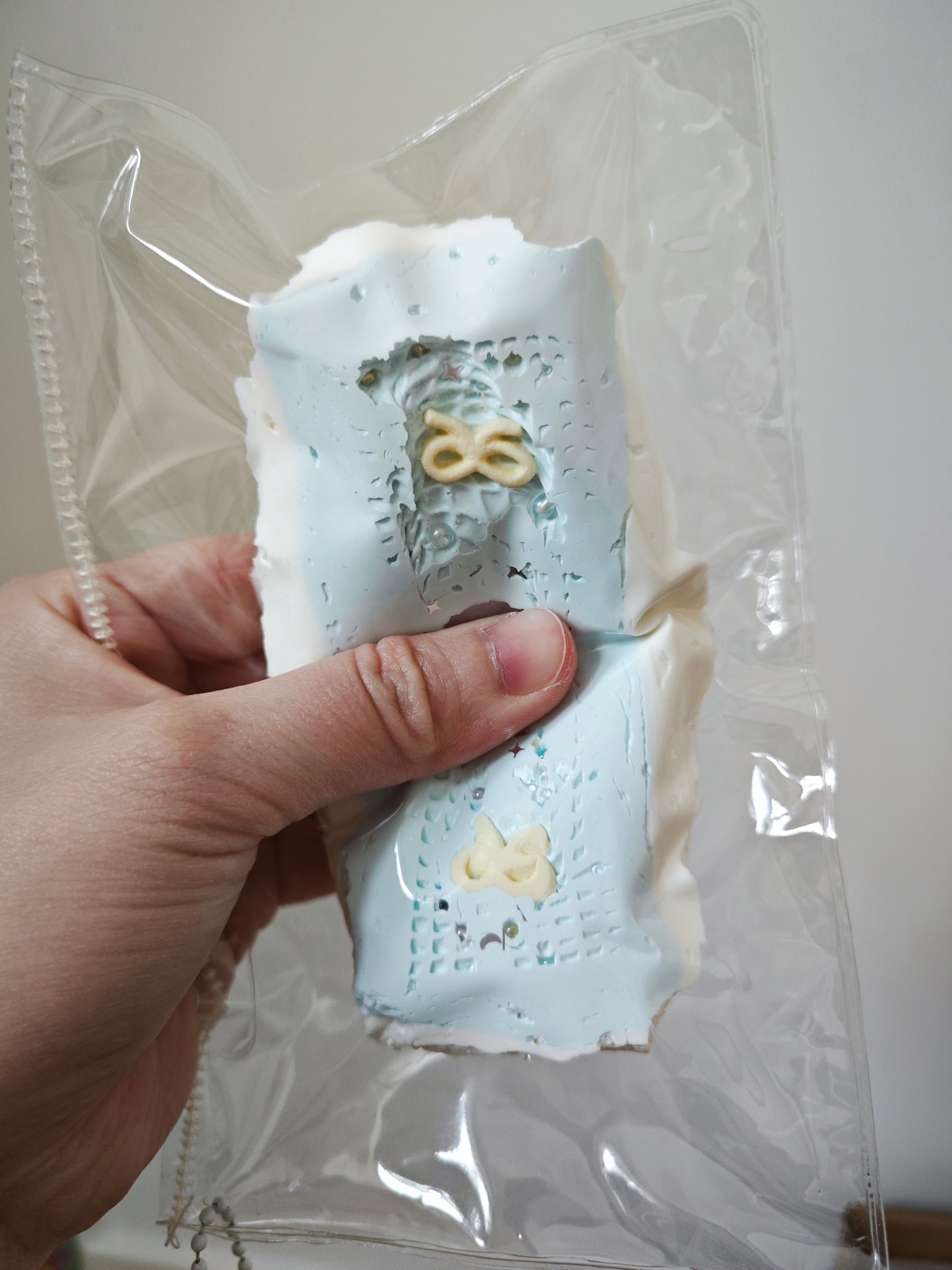 A hand holding a clear plastic bag containing two light blue textured rectangular items with small yellow decorative details, against a plain indoor background.