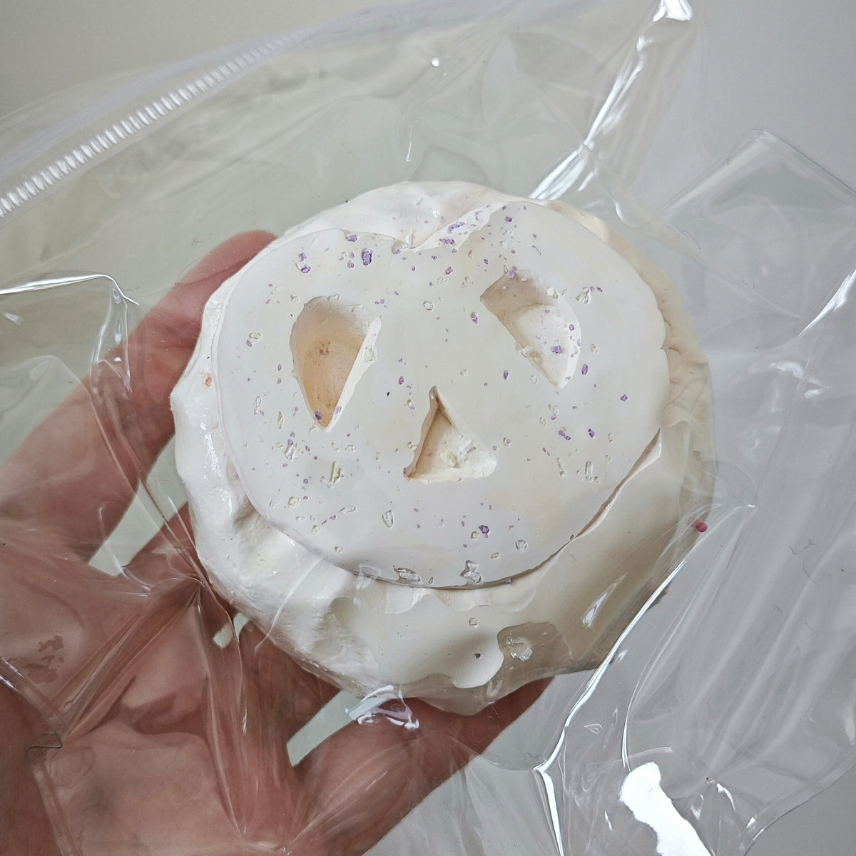 A hand holding a small white pumpkin-shaped item with a carved jack-o'-lantern face, wrapped in clear plastic packaging.